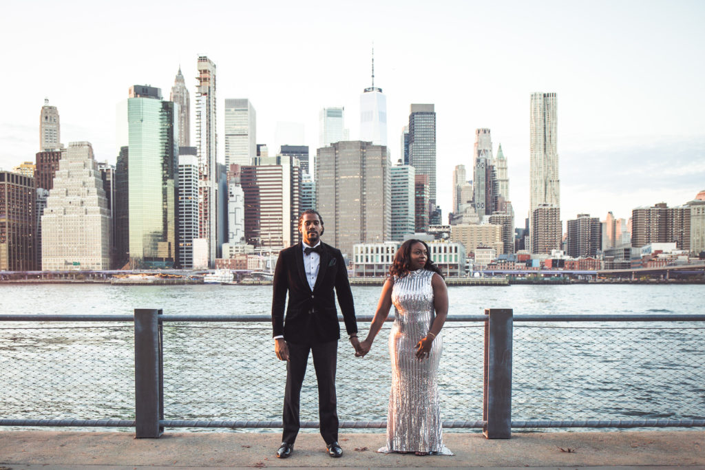 Man in Velvet Black Tuxedo holding the hand of Woman in Silver Sequin dress in front of New York City Skyline at Brooklyn Bridge Park, Pier 1.